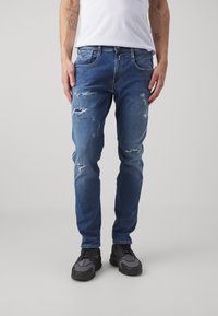Jeans de mezclilla azul desgastados con parches y bordes deshilachados, de corte slim. Combinados con zapatillas negras, con detalles en gris y textura prominente.