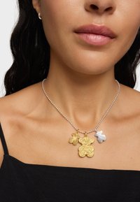 Collana con catena di perline d'argento che presenta tre pendenti: un orsetto di oro testurizzato, un fiore dorato e una forma geometrica bianca.