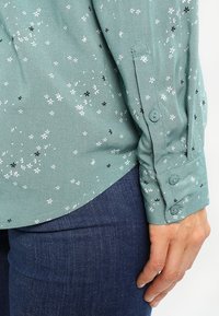 Blusa floreale verde con piccoli fiori bianchi e neri, tessuto liscio, polsini abbottonati, orlo leggermente curvato, abbinata a jeans blu.