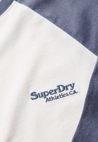 T-shirt met een witte body en donkerblauwe mouwen. Heeft "SuperDry Athletics.CA" geborduurd in lichtblauw. Zachte stoftextuur.