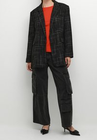 Sort mønstret blazer med et ternet design over en rød top, parret med sorte cargo-bukser med flere lommer og sorte spidse hæle.
