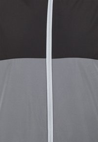Casaco corta-vento com uma parte superior preta e uma parte inferior cinza, com uma textura suave e um fecho central com zíper.