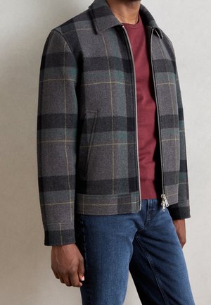 Mężczyzna w szarej i czarnej kratowanej kurtce na zamek, na bordo swetrze i niebieskich dżinsach, stojący na jednolitym tle.