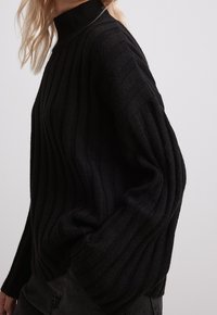 Pull en maille côtelée noire avec col montant, manches larges et coupe décontractée. La texture est lisse avec des motifs verticaux en relief.