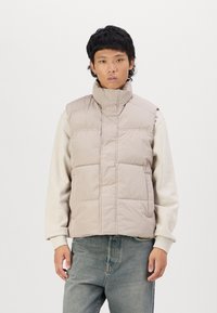 Puffer vest in lichtbeige, met een hoge kraag, gewatteerd ontwerp en een opening aan de voorkant. Draag het over een crèmekleurige sweatshirt en gecombineerd met blauwe jeans.