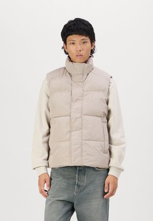 Puffervest i lys beige, med høj krave, quiltet design og frontåbning. Båret over en cremefarvet sweatshirt og parret med blå jeans.