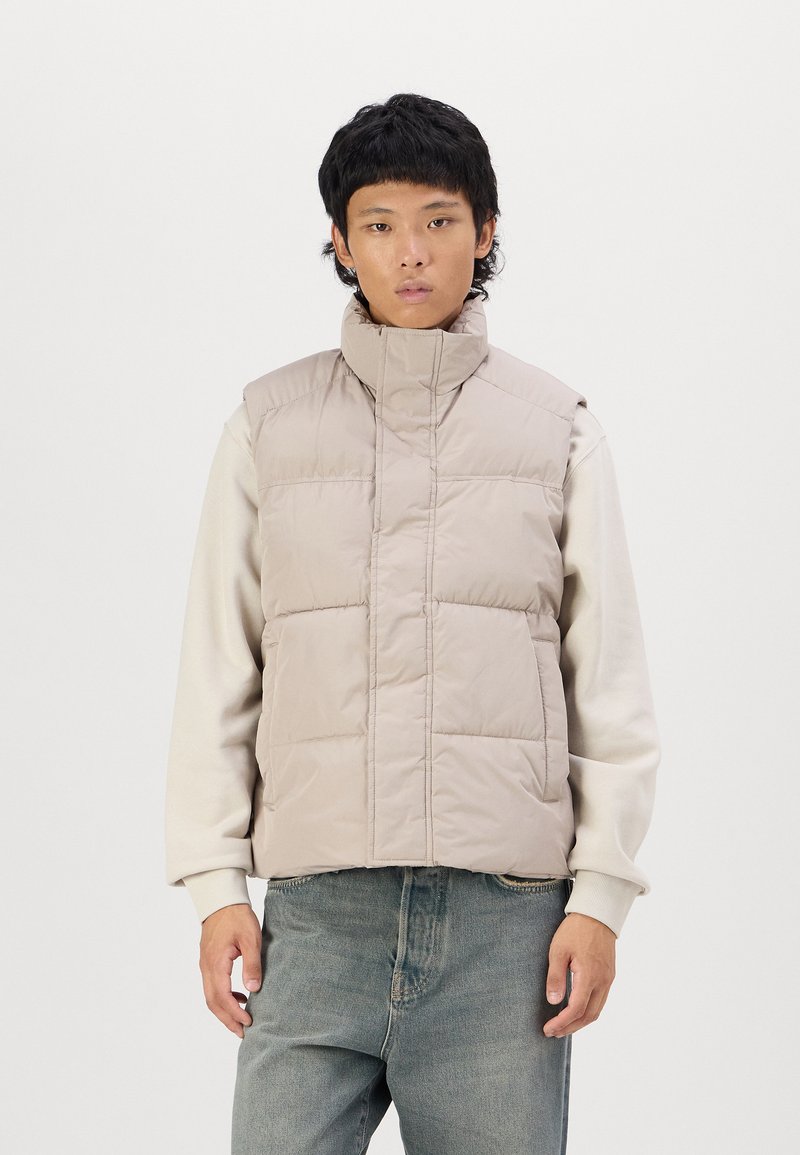 Puffer vest in lichtbeige, met een hoge kraag, gewatteerd ontwerp en een opening aan de voorkant. Draag het over een crèmekleurige sweatshirt en gecombineerd met blauwe jeans.