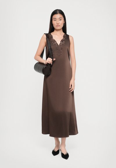 Rochie maxi fără mâneci, de culoare maro, cu decolteu în formă de V și detalii din dantelă. Modelul ține o geantă rotundă de culoare neagră, asortată cu pantofi eleganți negri cu vârf ascuțit.