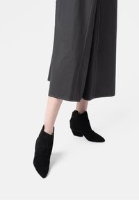 Czarne zamszowe botki do kostki z szpiczastymi noskami i niskimi obcasami, z dekoracyjnymi stężeńkami po bokach i zamkiem błyskawicznym z tyłu.