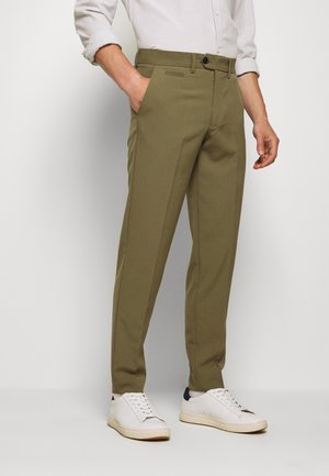 Mężczyzna ubrany w oliwkowozielone, dopasowane spodnie z przednimi kieszeniami, w białej koszuli z długimi rękawami i białych trampkach, stojący na jednolitym tle.