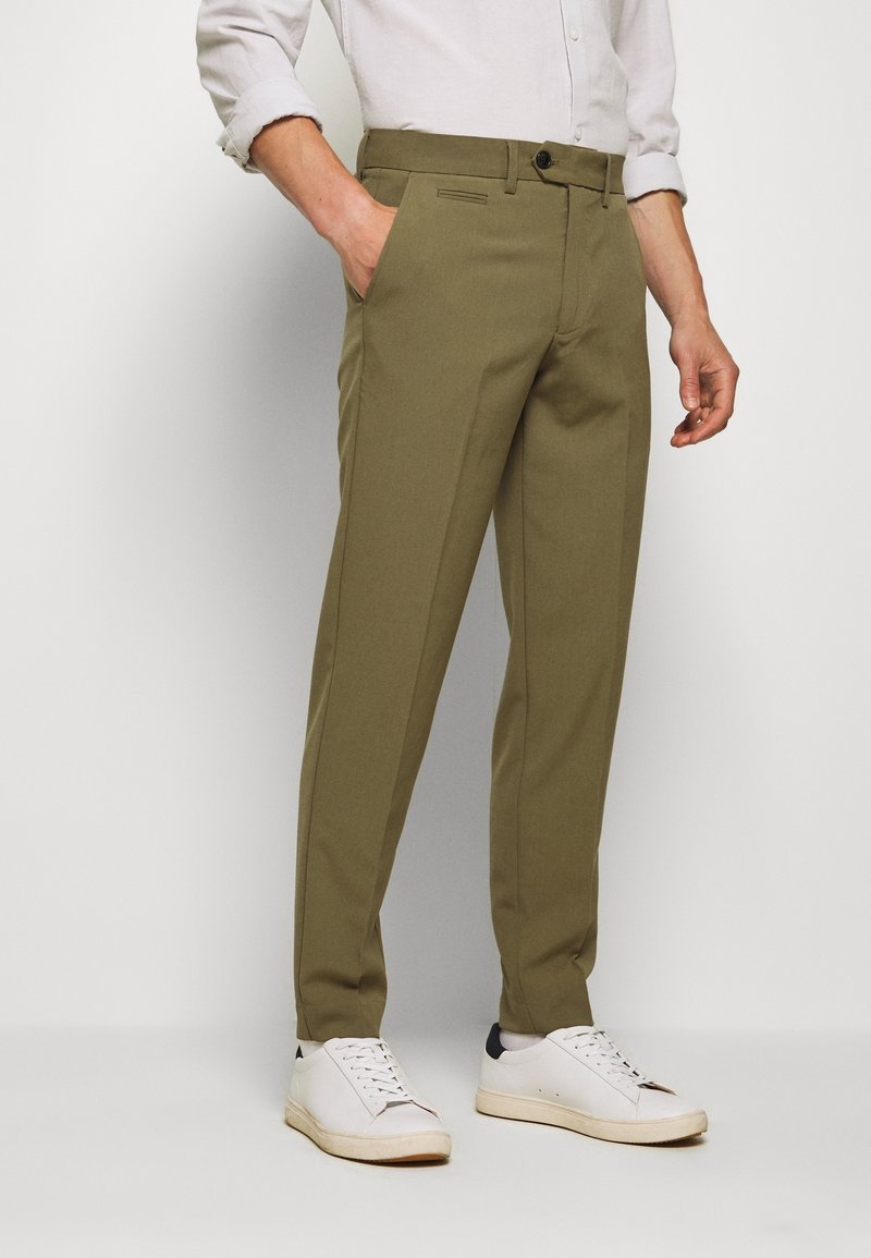 Homme portant un pantalon sur mesure vert olive avec des poches frontales, associé à une chemise blanche à manches longues et des baskets blanches, debout devant un fond uni.