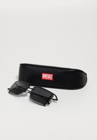 Black rectangular sunglasses with tinted lenses and metal accents, resting next to a black cylindrical case with a red Diesel logo.
