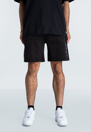Pantalones cortos de algodón negros con un corte recto, con un diseño lateral sutil, combinados con zapatillas blancas y una camiseta negra suelta.