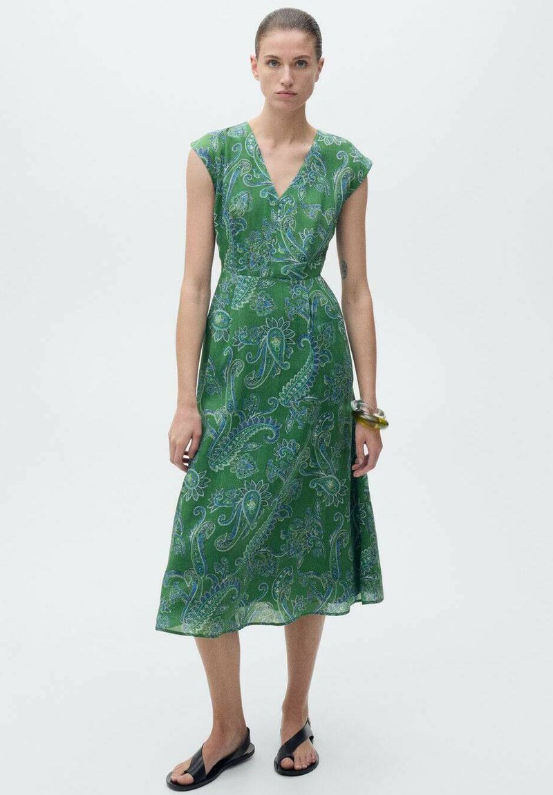 Grünes Paisleymuster-Kleid mit V-Ausschnitt, kurzen Ärmeln und taillierter Taille, das in Midi-Länge fließt. Getragen mit schwarzen Sandalen und einem gelben Armband.