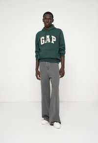 Zielona bluza z kapturem z logo "GAP" w kremowym kolorze, zestawiona z szerokimi szarymi dżinsami i białymi trampkami. Prosty, casualowy design, materiał bawełniany.