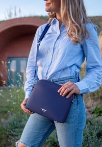 Navyfarbene Leder-Crossbody-Tasche mit glatter Textur, rechteckiger Form, silbernen Beschlägen und einem Logo auf der Vorderseite, gehalten gegen blaue Jeans.