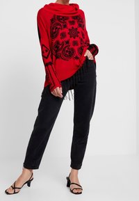 Rode trui met zwart, ingewikkeld bloem- en geometrisch patroon, off-shoulder col met druipende halslijn en franjes aan de zoom; gecombineerd met zwarte broek en hakken.