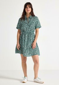 Grøn blomstrete kjole med korte ærmer, krave, knaplukning, indsnævret talje og udvidet nederdel. Bæres med hvide sneakers.