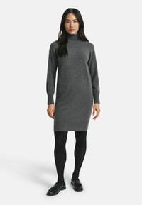 Abito a collo alto grigio realizzato in un morbido tessuto a maglia, design aderente, lunghezza al ginocchio con polsini e fondo a coste, abbinato a collant neri e mocassini.