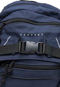 Mėlynas audinio kuprinė su keliais juodais užtrauktukais, plastiku uždarymu, tekstūruota paviršiumi ir baltu logotipu "FORVERT" priekyje.