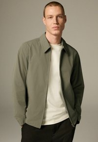 Lys grå zip-up jakke lavet af et blødt stof med spids krave. Båret over en ensfarvet hvid t-shirt, parret med sorte bukser.