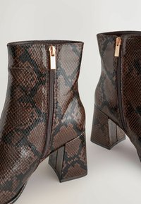 Brūni mākslīgās ādas potīšu zābaki ar rakstainu čūsku tekstūru, ar sānu rāvējslēdzēju un masīvu, kvadrātveida papēdi.