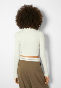 Bílý žebrovaný crop top s dlouhými rukávy, s vysokým límcem. Nosit s vysokými hnědými kalhotami s bílým pruhovaným pasem.