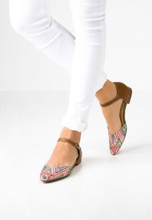 Pieds portant des chaussures marron à petits talons avec des bouts à motifs multicolores et des brides autour de la cheville, associées à un pantalon blanc retroussé sur un fond blanc.