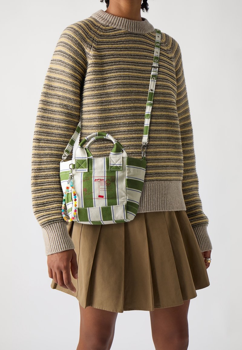 Personne portant un pull en maille rayé et une jupe plissée tenant un petit sac à main rayé vert et blanc avec un porte-clés en perles.