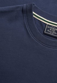 Marineblauwe crewneck-trui met geribbelde kraag en een Superdry & Co merklabel dat aan de binnenkant van de hals is genaaid.