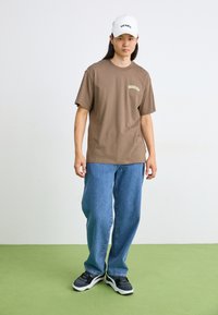 Barna pamut póló "DICKIES" logóval, kék laza farmernadrág, szürke és fekete sportcipő, fehér baseball sapka logóval. Egyszerű, hétköznapi ruházat.
