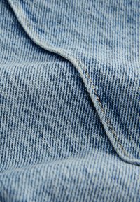 Denim v světle modré barvě s klasickým keprovým vzorem. Obsahuje viditelné švy v kontrastní béžové barvě a má jemný, texturovaný povrch.