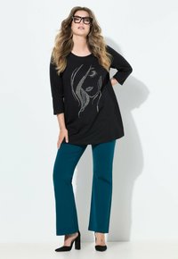 Túnica negra con un diseño de cara de strass, combinada con pantalones en tono azul verdoso. La modelo lleva puestos unos tacones negros y gafas de gran tamaño. Se observan texturas suaves.