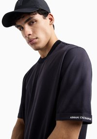 T-shirt nera a maniche corte con colletto tondo, dotata di un colletto a costine e logo "AR MANI EXCHANGE" ricamato sul polsino. Tessuto morbido.