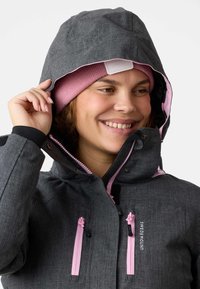 Graue wasserdichte Jacke mit strukturiertem Finish, pinken Reißverschlüssen und Kapuze. Darunter ein pinker Beanie, der den Kontrast der Farben betont.