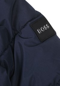 Sezione in giacca puffa blu navy con tessuto liscio e lucido; presenta una toppa quadrata nera con il logo "BOSS" in lettere argento.