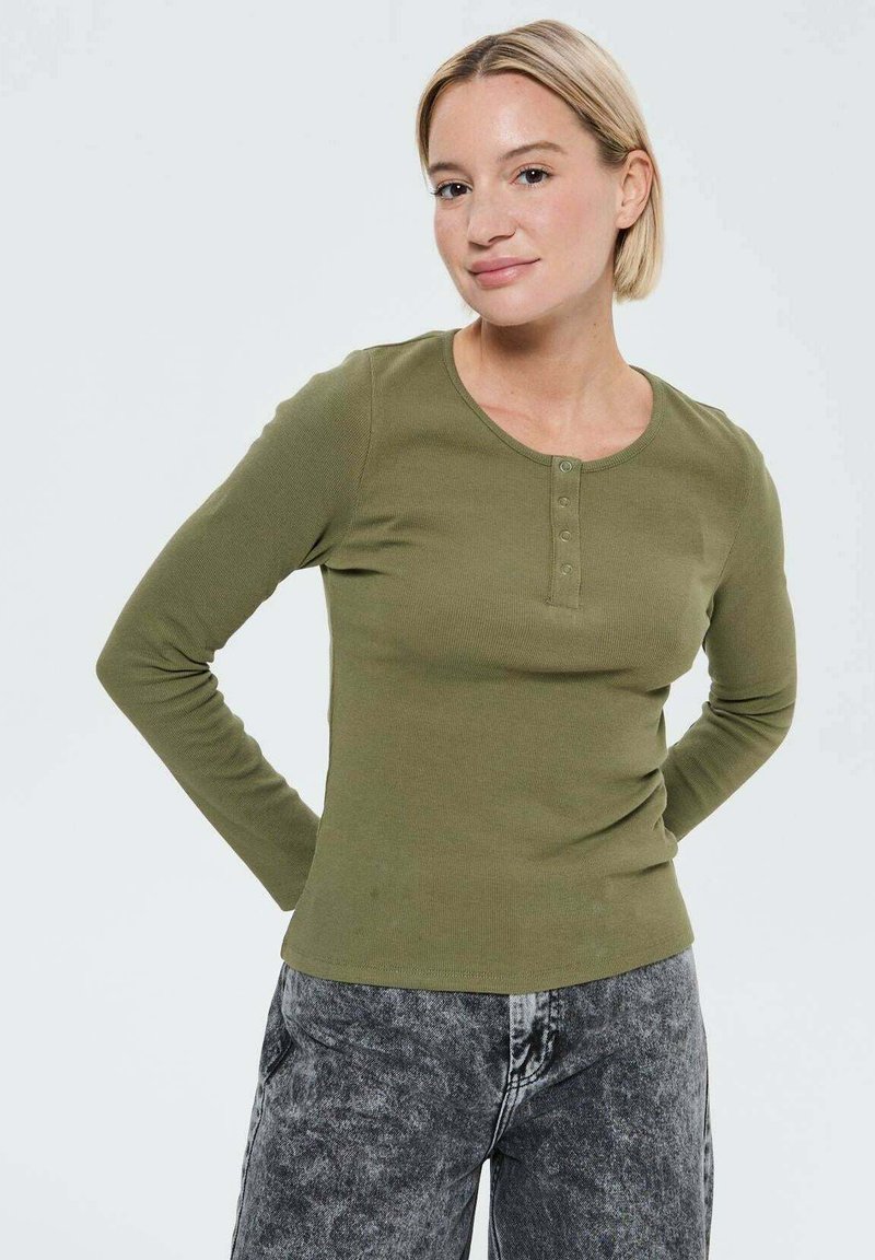 Haut à manches longues vert olive avec un col rond et une patte de boutonnage à cinq boutons. Texture côtelée douce. Associé à un jean en denim gris.