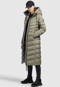Padded long coat in olive green with a hood, zip closure, side pockets, and elastic drawstrings. Paired with black jeans and sneakers.