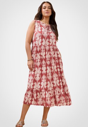 Kvinde iført ærmeløs, ankel-lang rød kjole med hvidt blomsterprint, kombineret med flade sandaler, stående mod en ensfarvet baggrund.