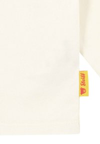 Langarmshirt aus Baumwolle in hellem Creme, mit einem gelben Etikett mit einem Bärenlogo und "Steiff"-Markierung auf der linken Seite.