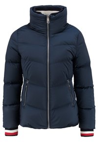 Veste matelassée bleu marine avec col haut, fermeture éclair frontale, poches latérales zippées, et poignets en tricot rayés blanc, bleu marine et rouge.