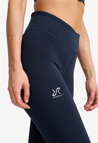 Närbild av en person som bär marinblå högmidjade leggings med RVRC-logotyp på låret, händerna avslappnat vid sidorna.