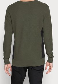 Felpa verde a maniche lunghe con texture, caratterizzata da un design a coste, pannelli laterali neri e polsini morbidi. Vista posteriore, con branding minimale.