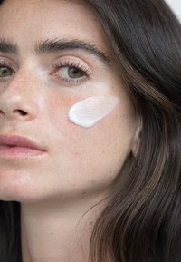 Fuktighetskrem smurt på lys hud, som viser en glatt, hvit tekstur. Modellen har mørkt hår og synlige fregner i ansiktet.