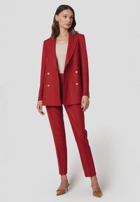 Czerwony garnitur szyty na miarę z podwójnym zapięciem i wąskimi spodniami. Żakiet ma złote guziki. Noszony z beżową bluzką i beżowymi szpilkami.