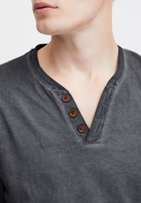 Graues Baumwollhemd mit V-Ausschnitt, drei braunen Holzknöpfen und strukturiertem Finish, das ein lässiges Design und weichen Stoff zeigt.