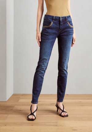 Femme portant un jean slim bleu foncé et des sandales à talons noires, debout sur un sol en bois devant un mur uni.