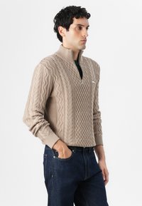 Maglione in maglia beige con colletto alto e zip a quarto. Presenta motivi testurizzati e polsini a costine. Abbinato a jeans di denim scuro.