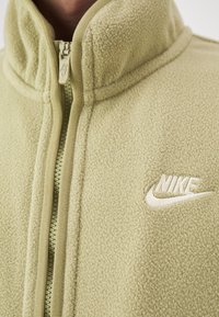 Fleece zip-up jacka i ljusgrön. Har en hög krage, texturerad tyg och broderad Nike-logotyp på vänster bröst. Dragkedjedetalj synlig.