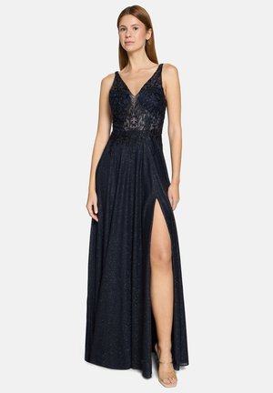 Woman wearing a sleeveless navy blue evening gown with sheer floral embroidered bodice and thigh-high slit, standing with neutral expression.